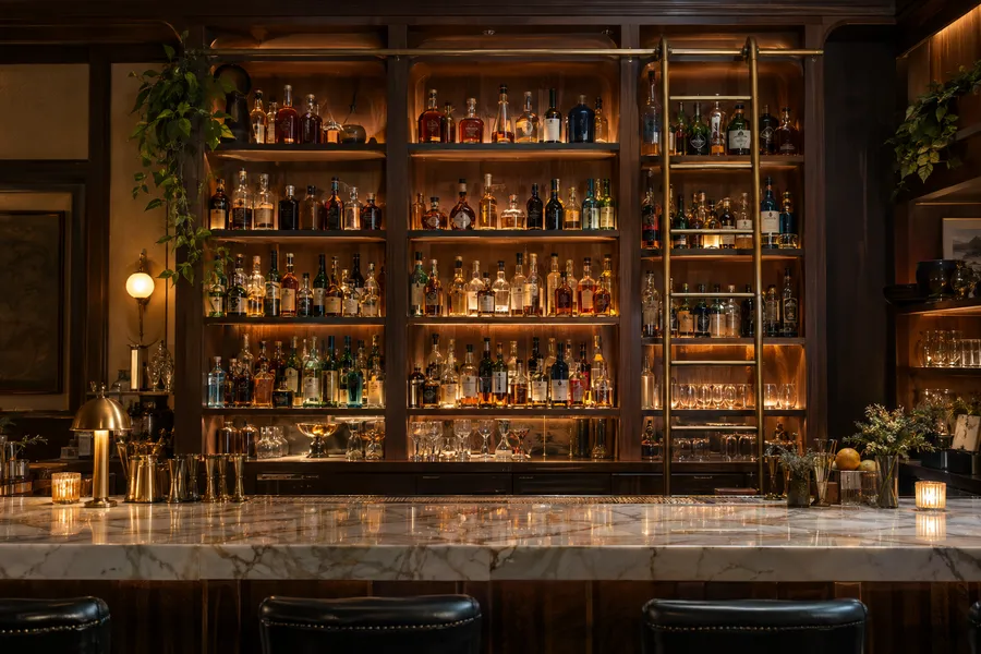 Concept image of a custom back bar with lit walnut shelving, a rolling brass ladder and a marble bar top.