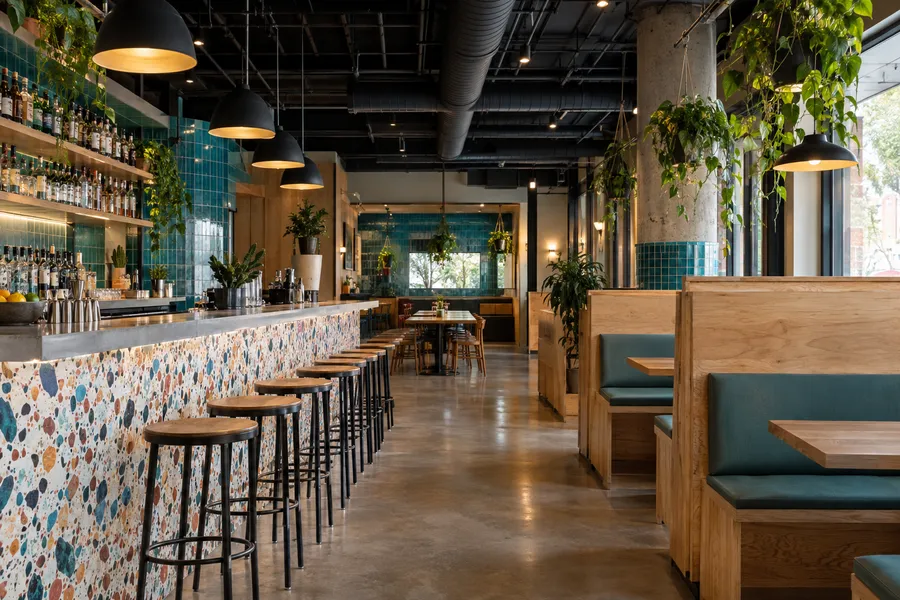 Concept image of a modern cantina with a terrazzo bar front, turquoise tile, plywood booths and hanging plants.