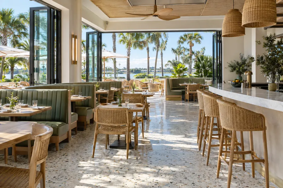 Concept image of a bright Orlando restaurant with sage banquettes, rattan chairs, terrazzo floor, a curved bar and a palm-lined patio.