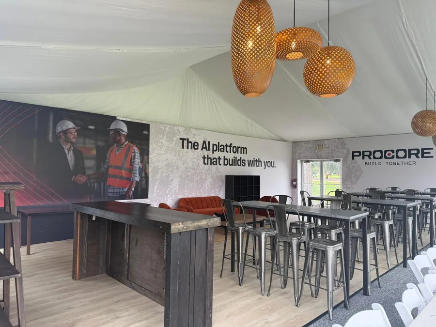 A long steel-and-timber service bar inside a tented pavilion, backed by a full-height printed brand mural and lit by woven pendant shades.