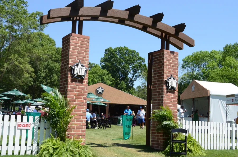 A brick-piered timber arch with branded roundels frames a white picket entry path onto a course, with guests entering beyond.