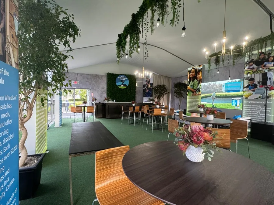 A lounge on turf flooring with a curved timber table, printed banner walls, trailing greenery overhead, and a bar counter behind.