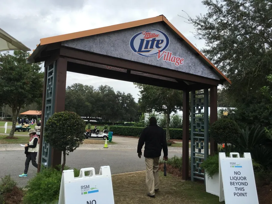 A timber gateway structure with a large branded panel above the opening, marking the entry to a spectator village on a course.