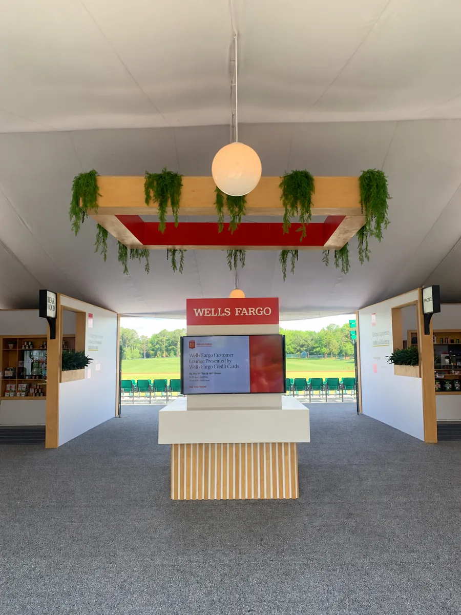 A Wells Fargo tent corridor with a timber greenery canopy, globe pendant, slatted pedestal and branded screen.