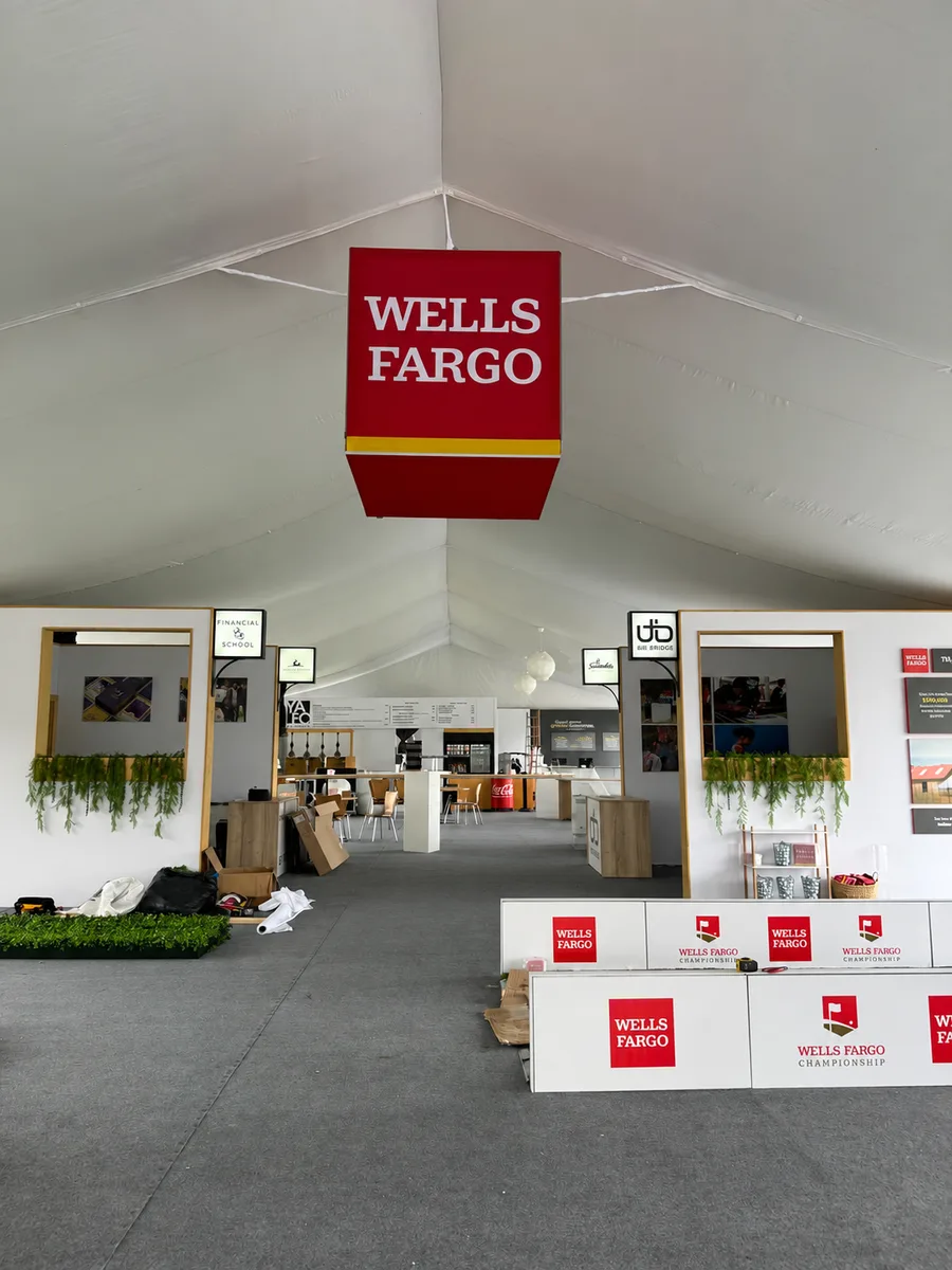 A Wells Fargo hospitality tent during setup, with a hanging branded cube, framed wall panels, greenery and branded counters.