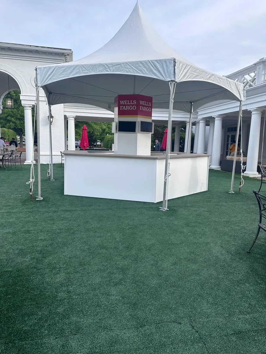 A white branded bar counter under a high-peak tent on turf at a tournament hospitality venue.