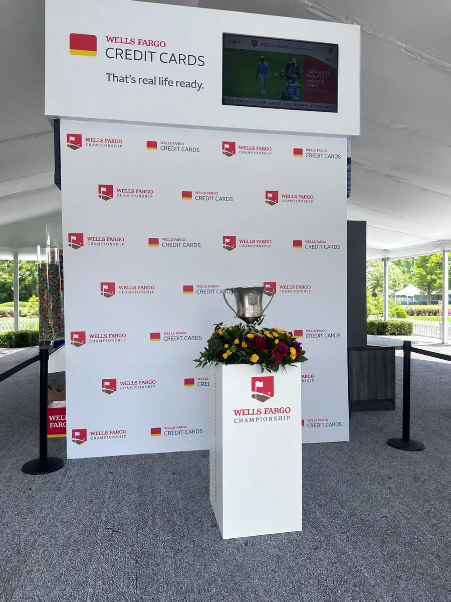 A Wells Fargo Credit Cards step-and-repeat backdrop with a trophy on a branded pedestal inside a hospitality tent.